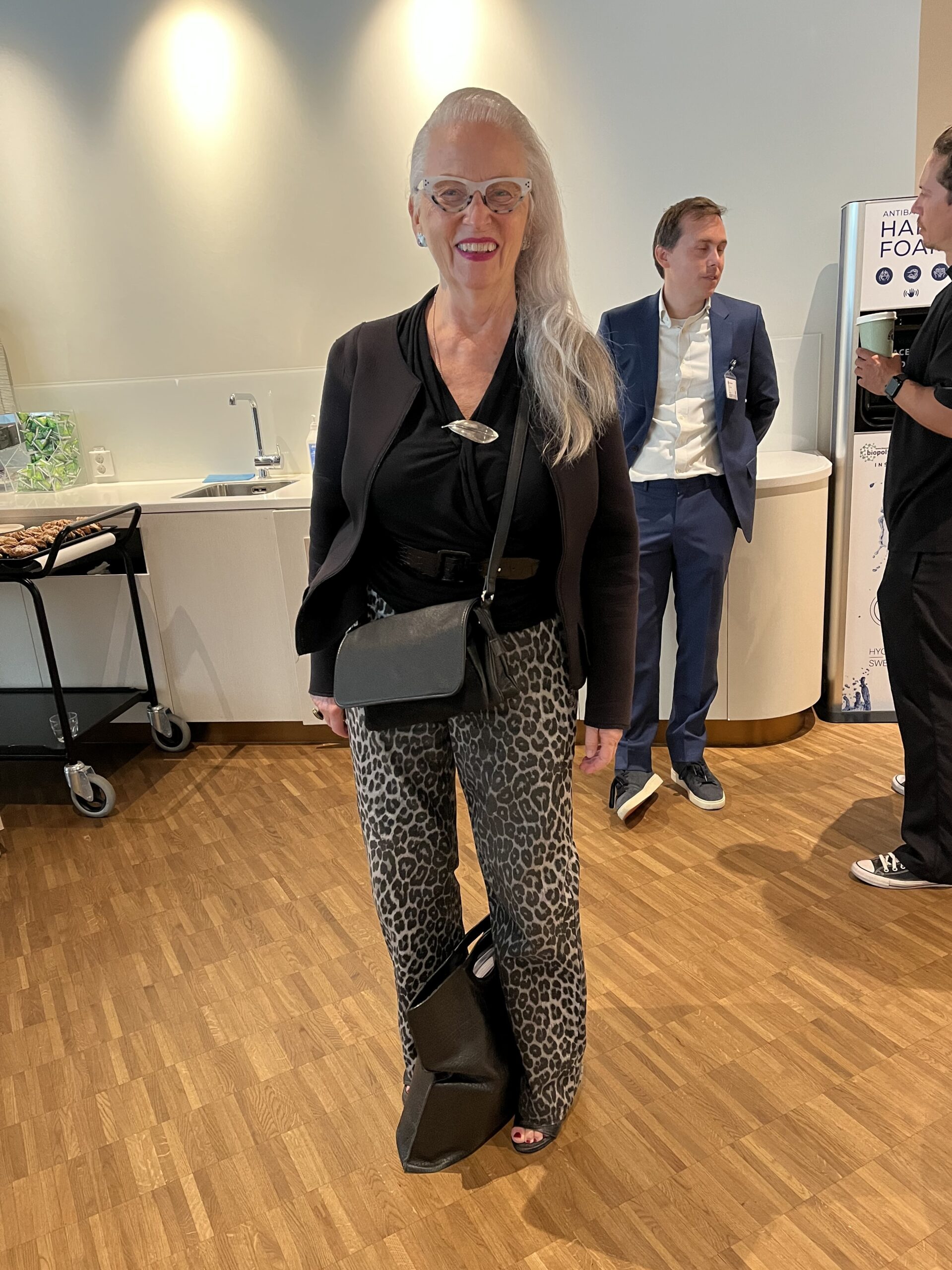 Kvinna med silverfärgat hår, glasögon och leopardmönstrade byxor står i ett modernt konferensrum.