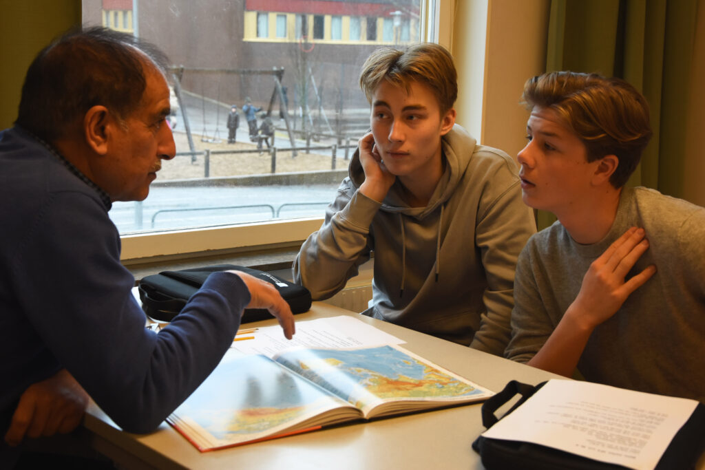 Vuxen man och två tonårspojkar i fokuserat samtal över en uppslagen atlas i klassrumsmiljö, del av språkutbytesprojektet Världen finns här.