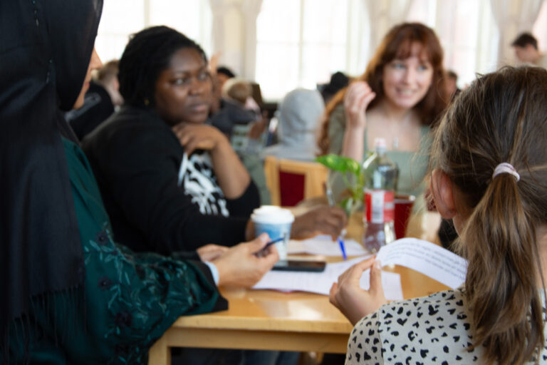 Vuxna och elever i gruppsamtal vid bord i skolmatsal under språkutbyte.
